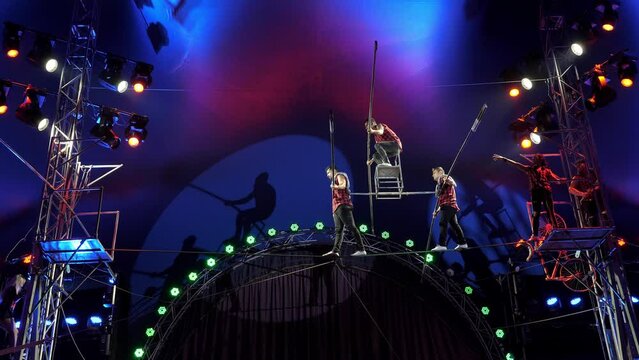 Aerial acrobats perform a difficult trick on a tightrope, two artists hold a rope with a chair on which a tightrope walker stands. Concert lighting with multi colored spotlights at a circus show.