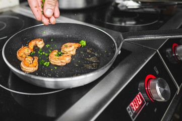 A close-up of a hand seasoning shrimps in a pan on an induction stove, highlighting a cooking moment