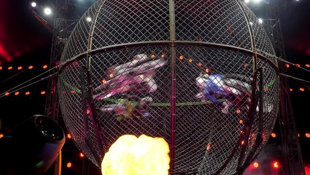 Four brave acrobats ride motorcycles in a large metal round cage in a circus with colorful lighting. Dangerous stunts while performing on a motorcycle. Acrobats on motorcycles. A circus show.