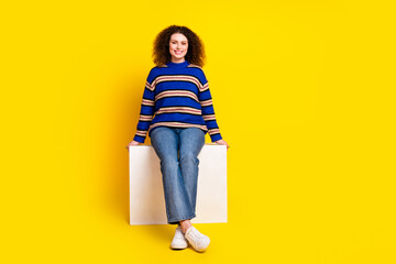 Full size photo of satisfied positive woman wear knit sweater jeans sit on white cube enjoy break...