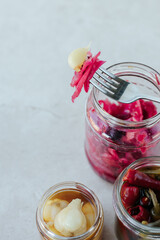Glass jars with fermented cabbage, garlic and pepper stand on a light stone background. On top of the jar lies a fork with cabbage and garlic