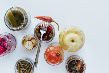 Glass jars with various fermented vegetables stand on a light background. There is a fork with chopped pepper at the top. shot from above