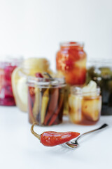 Glass jars with various fermented vegetables stand on a light background. in the foreground lies a fork with chopped pepper.