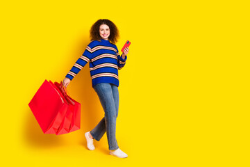 Full length photo of girlish woman wear knit sweater hold shopping bags smartphone go empty space...