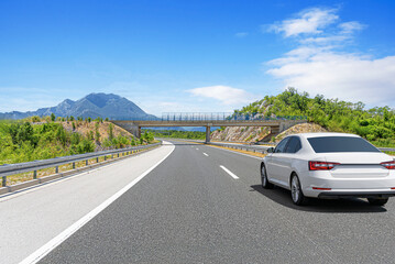 A white car drives along the highway against the backdrop of rocky mountains on a sunny day.