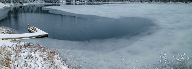 Panoramic view of a small lake with a fishing dock as the water in the lake is freezing into ice....