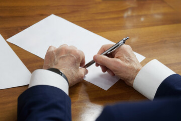 Elderly man sits at a table with documents and holds a metal fountain pen in his wrinkled hands....