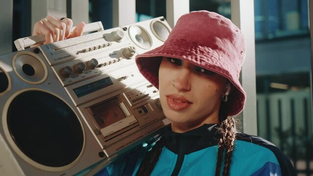 Creative young woman hip-hop dance performer posing standing with ghettoblaster on her shoulder in city downtown, retro music radio boombox, high-rise skyscrapers on background, 80s 90s urban style