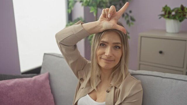 Young blonde woman sitting on sofa making fun of people with fingers on forehead doing loser gesture mocking and insulting. at home