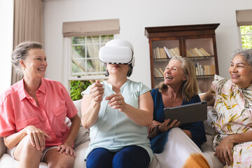 Diverse senior female friends trying on VR headset at home, laughing together