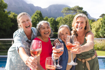Diverse senior female friends holding wine glasses, laughing outdoors