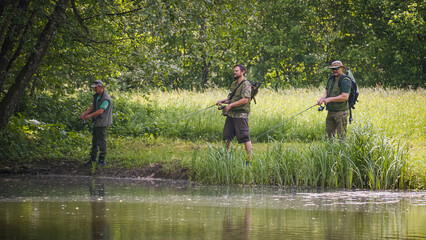 Three sports fishermen standing on the river bank, under a beautiful tree, swinging their fishing...
