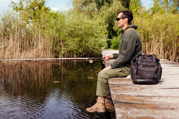 Young man fishing wearing military uniform. Guy sitting on bridge across river holding rod....