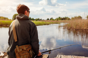 Back view of man fishing wearing military uniform. Fisherman sitting on bridge across river holding rod. Recreation