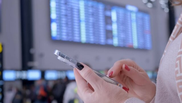 Bottom side view of female passenger without face standing near arrival departure board at airport terminal, tapping on smartphone screen, leaning hands on luggage bag handle. Mobile check-in concept