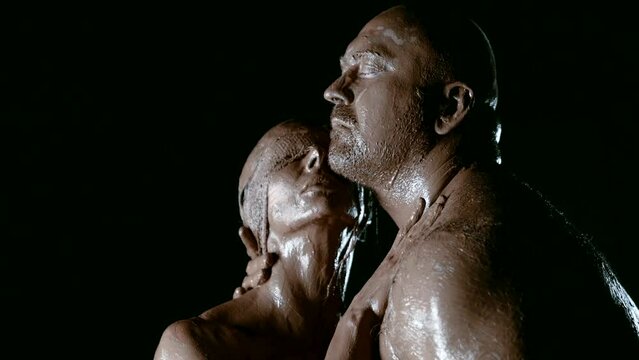 In the darkness of the night, a man with a strong physique and his wife, covered in clay, reveal their passion and unbreakable bond on a black background.