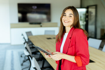 Smiling professional woman in red blazer holding tablet in modern conference room during daytime