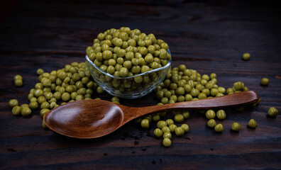 A glass cup with green canned peas stands on a wooden surface. A wooden spoon lies next to a cup with green peas.