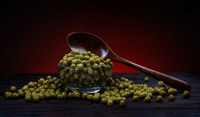 A glass cup with green canned peas stands on a wooden surface. A wooden spoon lies on a cup with canned green peas. Beautifully illuminated background.