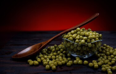 A glass cup with green canned peas stands on a wooden surface. A wooden spoon lies on a cup with canned green peas. Beautifully illuminated background.