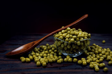 A glass cup with green canned peas stands on a wooden surface. A wooden spoon lies on a cup with canned green peas. Beautifully illuminated background.