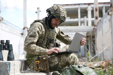 Military mission. Soldier in uniform using laptop near abandoned building outside