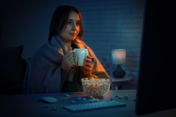 Beautiful young woman under blanket with popcorn and cup of drink watching film on computer at...