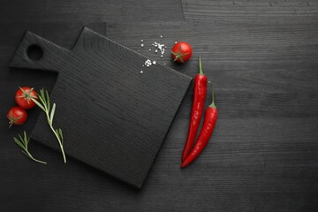 Cutting board, rosemary, chili peppers and tomatoes on black wooden table, flat lay. Space for text