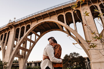 LGBTQ+ engaged couple outdoors cuddling under a bridge