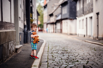 A young boy holding a teddy bear stands on a cobblestone street in a charming residential area. He...