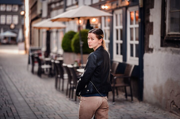 A young woman wearing a black leather jacket looks back over her shoulder while walking on a...