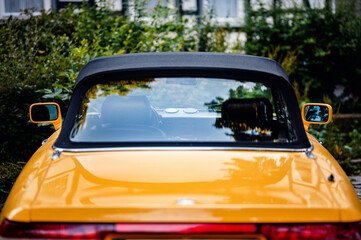 Back view of a vintage yellow convertible car with a black soft top, parked outdoors near greenery,...