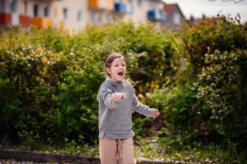 A young girl enjoying a spring day, holding out her hand to catch cherry blossom petals, surrounded by lush greenery and colorful buildings in the background.