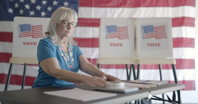 Three quarter angle, medium shot, elderly woman election worker checks over forms and stickers for voters at polling station. Flag and voting booths behind