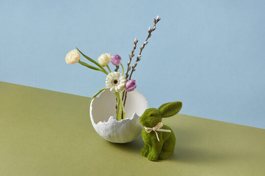 Easter toy next to vase in form of broken eggshell with spring flowers
