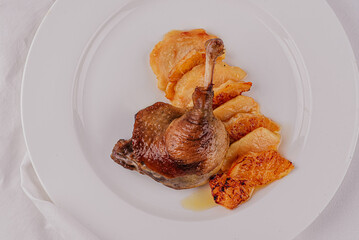 Rosy roasted duck leg with a crispy crust, grilled oranges and apple pieces, on a white plate and white tablecloth. Horizontally