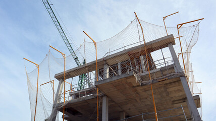Multi-story concrete building is under construction, featuring scaffolding and safety nets. Project is located outdoors on clear land with a blue sky overhead.