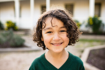 School age boy with dark curly hair smiles outside in front of California adobe in the fall
