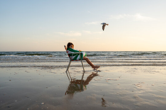 A landscape of a girl at the beach sitting on a beach chair.