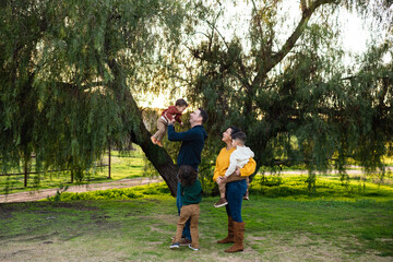 Lifestyle portrait of young family together in front of tree at sunset as dad lifts toddler into the air