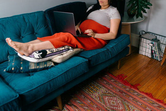 pregnant woman sitting with feet up on sofa  scrolling on  laptop