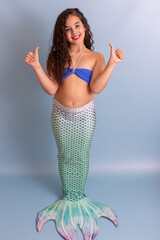 Beautiful happy girl in a mermaid costume stand on a blue background showing thumbs up.