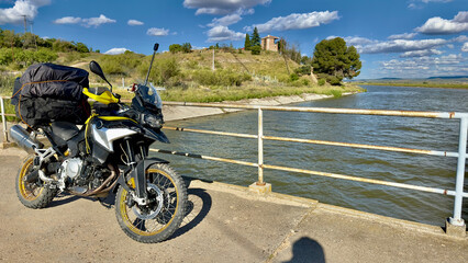 A fully equipped adventure motorcycle with off-road tires and full equipment parked next to a picturesque river on a sunny day. The bike is equipped with a large backpack and a robust exhaust system.  © Diego Schiochet