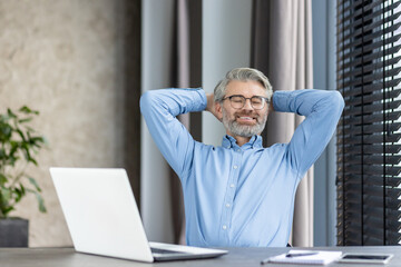 A content elderly man is happily relaxing with hands behind his head at his work desk
