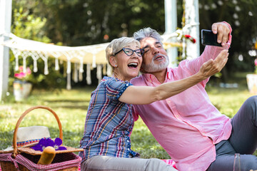 Modern elderly couple taking selfie on smartphone, having picnic and resting in garden at sunny day