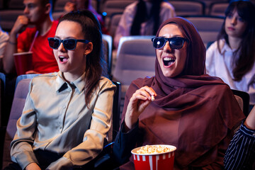 Hijab muslim woman watching a 3d movie in cinema with her friends. Eating popcorn and enjoying.