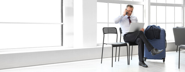 Mature businessman with laptop talking by mobile phone in hall of airport