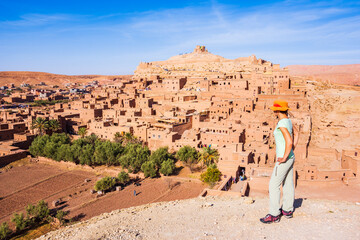 Young woman tourist in orange hat standing on viewpoint over Ksar Ait Ben Haddou, old Berber...
