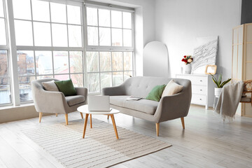 Interior of light living room with grey sofa, armchair and laptop on table