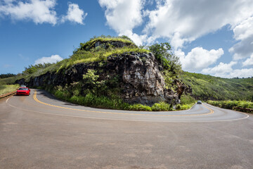Red car on reverse road curve on Maui around a rocky cliff with green vegetation, tropical vacation...